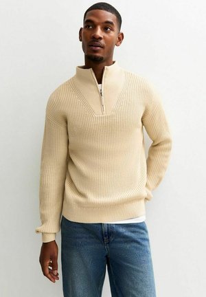 Beige gebreide pullover met een half-rits kraag, gekenmerkt door een textuurpatroon en geribde manchetten, gecombineerd met denim jeans.