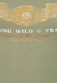 Zelená látka s bledým bílým textem "Riding Wild & Free", červeným kurzívou "Queen of the Road" a žlutými dekorativními křídly a emblémem nahoře.