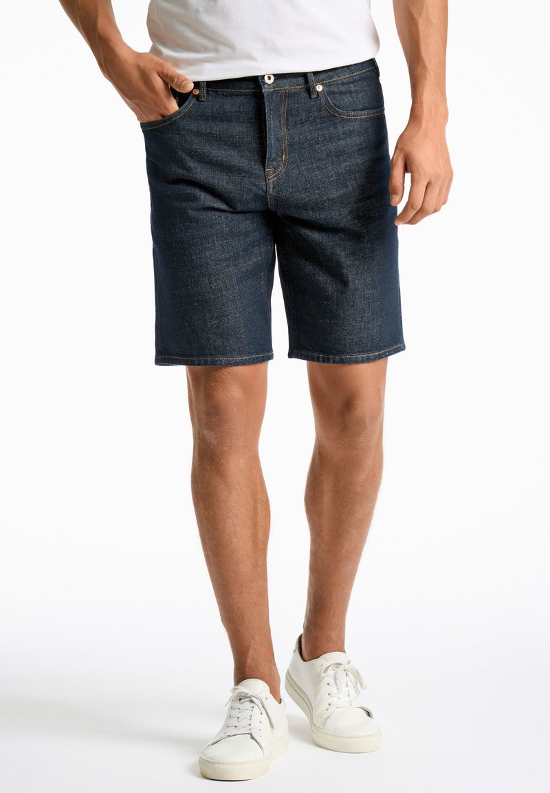 Dunkelblaue Denim-Shorts mit geradem Schnitt, ausgestattet mit fünf Taschen und kontrastierendem orangefarbenem Stich, kombiniert mit weißen Sneakers.