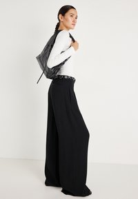 Haját fonatban viselő nő fehér, hosszú ujjú felsőt, széles lábú fekete nadrágot, szegecses övet hord, fekete, texturált válltáskát cipel, profilból áll.