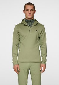 Oliivinvihreä vetoketjullinen huppari, jossa on huppu ja sivuvetoketjutaskut; sisältää teksturoidun kankaan ja mustan logon rinnassa; sileä pinta.