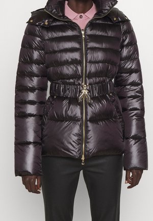Veste matelassée noire brillante avec quilting horizontal, fermeture éclair, taille ceinturée avec une boucle d'insecte dorée décorative, portée sur une chemise rose à col.