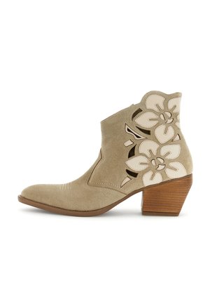 Bottine en daim beige avec talon bloc en bois et motifs floraux découpés sur le côté, présentant un bout pointu et des détails cousus.