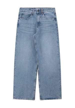 Femme aux longs cheveux foncés portant un t-shirt crème et un jean bleu à jambes larges, debout, une main couvrant son visage, souriante sur un fond beige.