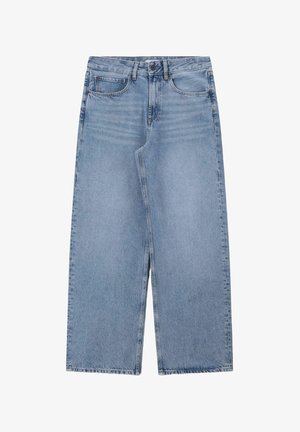 Femme aux longs cheveux foncés portant un t-shirt crème et un jean bleu à jambes larges, debout, une main couvrant son visage, souriante sur un fond beige.
