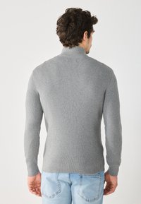 Suéter gris de punto acanalado con cuello alto y mangas largas, llevado sobre unos vaqueros azul claro. La parte de atrás presenta un diseño ajustado con tela texturizada.