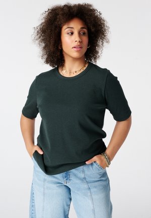 Vrouw met krullend haar die een donkergroen shirt met korte mouwen en lichtblauwe spijkerbroek draagt, met de handen in de zakken, kijkt naar de zijkant.