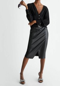 Donna in una blusa nera a scollo a V profondo e una gonna midi in ecopelle nera con una tasca grande, che tiene degli occhiali da sole in una mano.