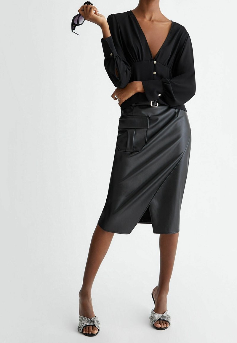 Donna in una blusa nera a scollo a V profondo e una gonna midi in ecopelle nera con una tasca grande, che tiene degli occhiali da sole in una mano.