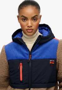 Hooded jack met blauwe corduroy en zwarte stofsecties, voorzien van oranje ritsen en een bruine fleece textuur op de mouwen. Logo patch op de borst.