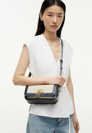 Mujer con cabello negro liso lleva una camiseta blanca sin mangas y vaqueros azul claro, sosteniendo un bolso bandolera beige y negro con logo dorado.