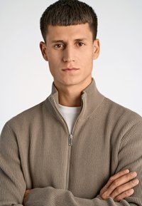 Maglione beige a coste con zip e colletto alto, indossato su una camicia bianca. La texture è morbida e lavorata a maglia, con un dettaglio della zip in argento.