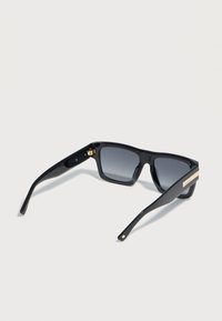 Gafas de sol rectangulares negras con lentes de degradado oscuro y detalle de marca dorada en las patillas, mostradas en una vista lateral ligeramente inclinada.