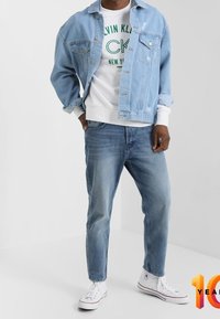 Lichtblauwe spijkerjas over een wit sweatshirt met groene merkletters, gecombineerd met lichtblauwe spijkerbroeken en witte sneakers.