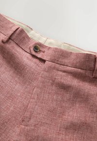 Gros plan sur un pantalon rose texturé avec fermeture à bouton, passants de ceinture et doublure intérieure beige de la ceinture sur un fond clair.