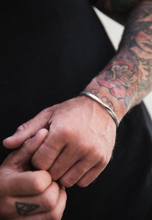 Silver cuff bracelet on a hand with intricate tattoos, featuring a smooth, polished surface and a simple, curved design.