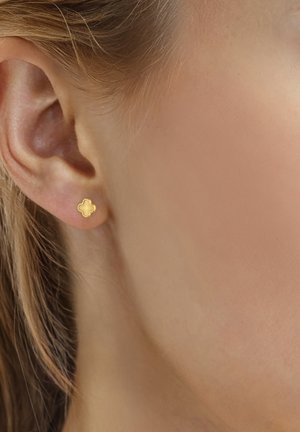 Primer plano de la oreja de una mujer que lleva un pequeño pendiente de botón en forma de trébol de cuatro hojas en oro, con detalles texturizados.