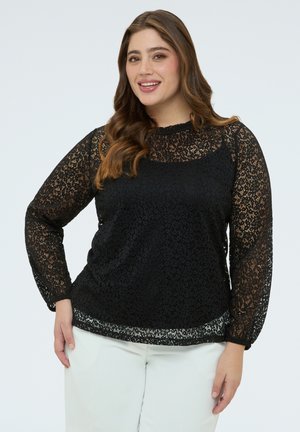 Donna sorridente con lunghi capelli castani che indossa una maglia nera in pizzo trasparente a maniche lunghe sopra una canottiera nera e pantaloni bianchi, su uno sfondo semplice.