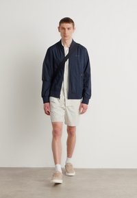 Marine bomberjack, wit gestreept shirt, lichtgekleurde shorts en beige sneakers. Model draagt een zwarte schoudertas en staat op een neutrale achtergrond.