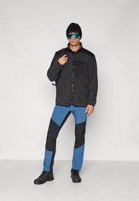 Fleece jacket in dark gray with black shoulder accents, paired with blue and black patterned pants. Accessories include a black beanie and sunglasses.