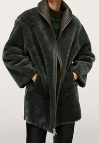 Korte donkergroene nepbonten jas met een hoge kraag, ritssluiting en zijzakken. Heeft een zachte textuur en een oversized pasvorm.