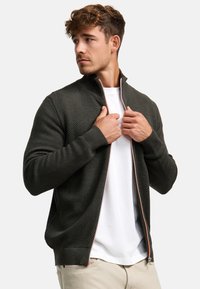 Mørkegrøn strikket zip-up jakke med tekstureret stof, ribbede manchetter og en kontrastfarvet lynlås. Bæres over en hvid T-shirt, hvor sidesømmene er synlige.