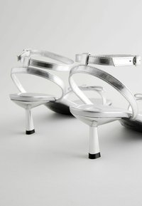 Silver high-heel sandals with a metallic finish, featuring thin straps and a tapered heel, accented with black detailing at the sole.