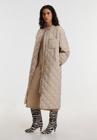 Beige gesteppter langer Mantel mit Reißverschluss, ausgestattet mit einer Brusttasche und einem gerippten Kragen, getragen über einem beigen Kleid und zebra-gestreiften Stiefeln.