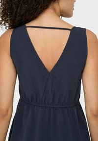 V-Ausschnittkleid aus marineblauem Stoff, mit gerafftem Taillenbereich, verstellbaren Trägern und offenem Rückendesign. Weicher Stoff mit dezentem Glanz.
