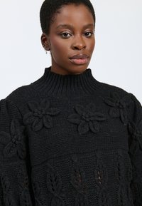 Pullover nero in maglia con un alto colletto alla mordacchia, caratterizzato da motivi floreali e fogliari, che evidenziano cuciture testurizzate e accenti dimensionali.