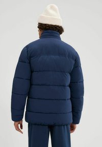 Chaqueta acolchada azul marino con costuras horizontales, cuello alto y puños elásticos, vista desde la espalda. Combinada con un gorro de lana color crema.