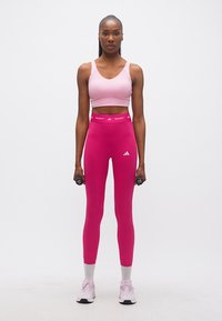 Γυναίκα με ροζ αθλητικό σουτιέν, φούξια κολάν και αθλητικά παπούτσια, όρθια και κρατώντας μαύρα βαράκια στα πλευρά της.