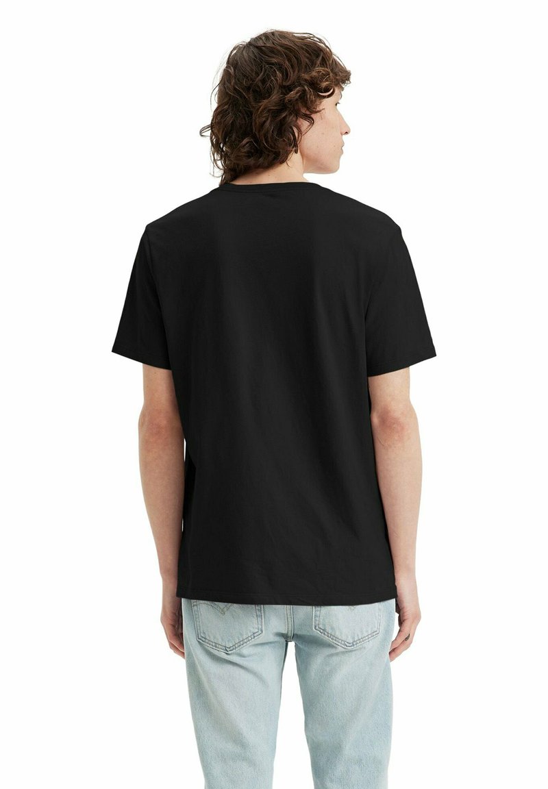 Junge Person mit lockigem Haar, von hinten gesehen, trägt ein schlichtes schwarzes kurzärmeliges T-Shirt und hellblaue Jeans vor weißem Hintergrund.