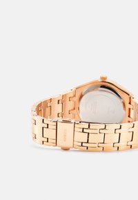 Orologio in acciaio inossidabile dorato con un design a bracciale a maglie. Presenta una cassa rotonda con un fondello trasparente e una corona rialzata sul lato.