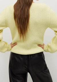 Pull en maille côtelée jaune clair avec manches larges, porté avec un pantalon en cuir noir doté de deux poches arrière. Le contraste de texture et de couleur est notable.