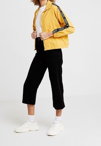 Osoba ubrana w żółtą kurtkę w czarne paski, czarne krótkie spodnie, białe trampki i białe skarpetki, stojąca na jednolitym tle.