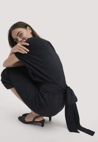 Eine Frau hockt in einem schwarzen, taillierten Overall und schwarzen Slingback-Absätzen, während sie mit einem sanften Lächeln in die Kamera schaut.