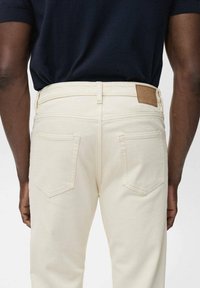 Pantaloni din denim bej deschis cu croială dreaptă, având buzunare pe spate și o etichetă din piele maro la talie. Textură netedă, design minimalist.