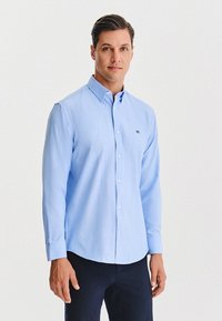 Jasnoniebieska, długosleeve, koszula zapinana na guziki, wykonana z bawełny, z klasycznym kołnierzykiem, przednia kieszeń z logo, slim fit, połączona z ciemnymi spodniami.