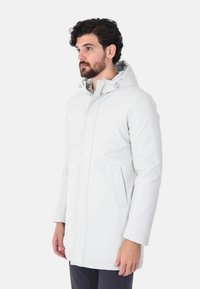 Parka lunga grigio chiaro con cappuccio removibile, chiusura con zip, due tasche laterali e una texture liscia e elegante.