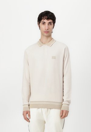 Langærmet beige polo med en struktureret overflade, ribbet krave og manchetter, prydet med et diskret logo på brystet og kontrasterende striber i kanten.