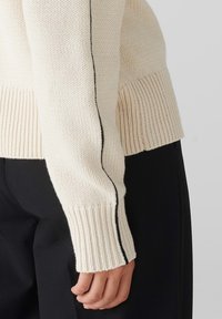 Cremfarbene Strickpullover mit gerippten Bündchen und Seitennähten, die einen schwarzen Streifen entlang der Länge aufweisen. Strukturiert, entspannte Passform.