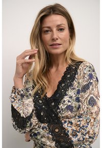 Blouse à motif floral en crème, avec des dentelles noires à bordure circulaire. Manches longues, coupe décontractée, motif floral multicolore.