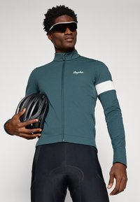 Chaqueta de ciclismo de manga larga en verde oscuro con un acento blanco en la manga y un cuello alto; el modelo sostiene un casco negro; pantalones cortos de ciclismo ajustados en negro.