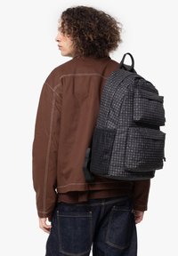 Mochila de cuadros en negro y gris con múltiples bolsillos, correas ajustables y una superficie texturizada, llevada en la espalda de una figura con chaqueta marrón.