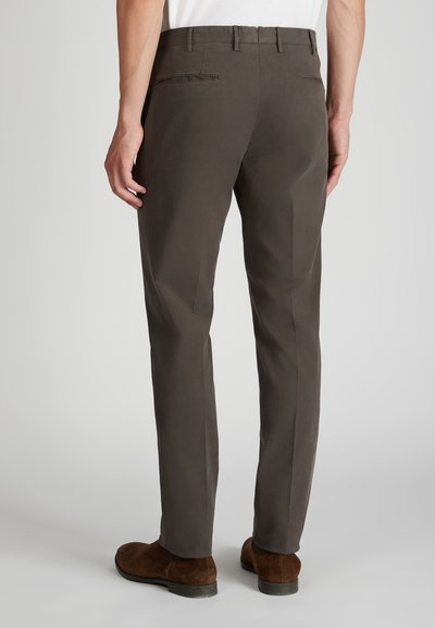 Hombre vistiendo pantalones de traje gris oscuro y zapatos de ante marrones, visto desde la espalda sobre un fondo claro y uniforme.