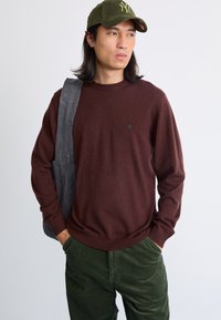Suéter marrón de punto con escote redondo, que presenta un pequeño logo negro. Combinado con pantalones de pana verde y un bolso de hombro gris oscuro.
