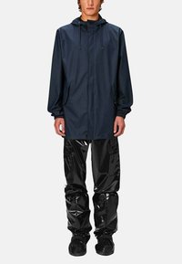 Veste imperméable de navy avec capuche et cordons, associée à un pantalon imperméable noir brillant et des chaussures noires à enfiler. Surfaces texturées visibles.
