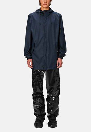 Giacca impermeabile blu navy con cappuccio e cordini, abbinata a pantaloni impermeabili neri lucidi e scarpe slip-on nere. Superfici texture visibili.