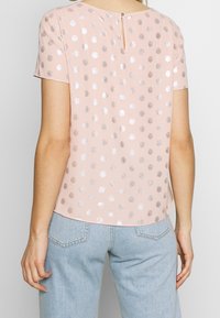 Blusa rosa claro con lunares plateados, manga corta, escote redondo y un detalle de abertura en la parte posterior. Tejido suave con una textura lisa.